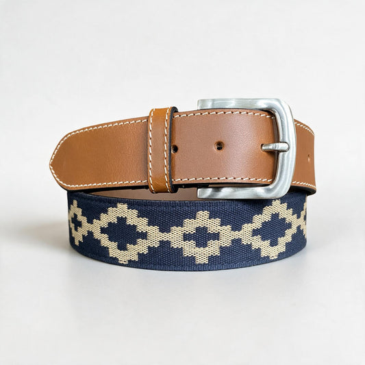 Cinturón Keltehue enrollado sobre fondo gris claro de estudio, cinta tejida con diseño guardapampa en azul marino y beige